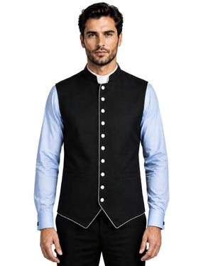 A man modeling a black waistcoat vest with white piping and white buttons, worn over a light blue dress shirt and black trousers, against a white background.
