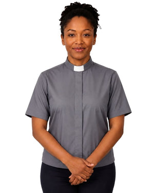 A woman wearing a short-sleeved gray clerical shirt with a white collar, standing against a plain white background.