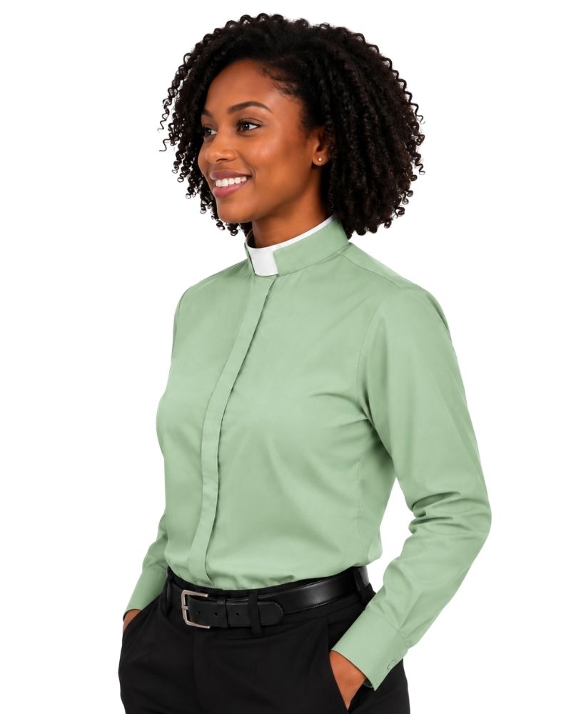 A smiling woman with curly black hair wearing a sage green long-sleeve clerical shirt with a white collar tab, paired with black trousers and a black belt, against a plain white background.