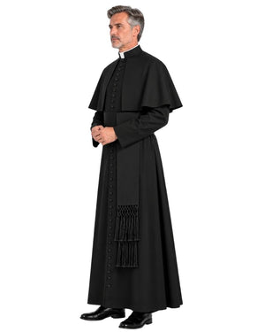 A man modeling a long black clerical cassock with a high collar, front buttons, wide sleeves, and a decorative tassel detail near the lower front, photographed against a plain white background.