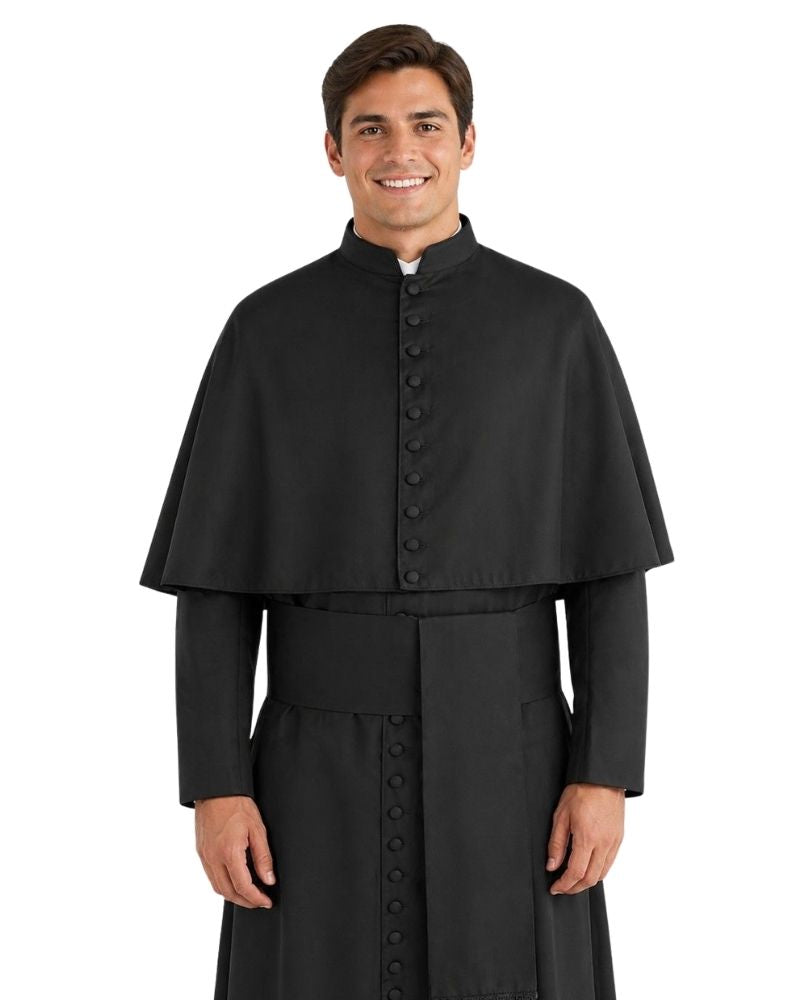 A man wearing a black clerical cape and matching black robe over a white collar, standing against a plain white background.