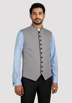 Man wearing a gray vest with black buttons over a light blue shirt with a white collar on a plain background