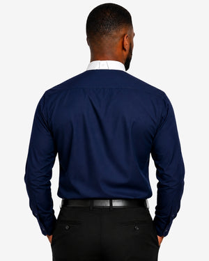 A man viewed from the back wearing a navy blue long-sleeve clerical shirt, a white collared undershirt, and black trousers against a light gray studio background.