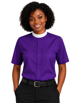 A smiling woman with curly black hair wearing a purple short-sleeve clerical shirt with a white mock neck collar, paired with black trousers and a black belt, against a plain white background.