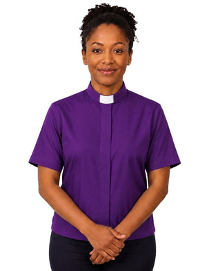 A woman wearing a purple short-sleeve clerical shirt with a white collar, standing against a plain white background.