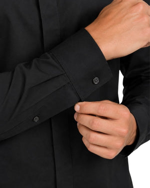 Close-up of a man wearing a black long-sleeve clerical shirt and buttoning the cuff with one hand while the other hand rests nearby.