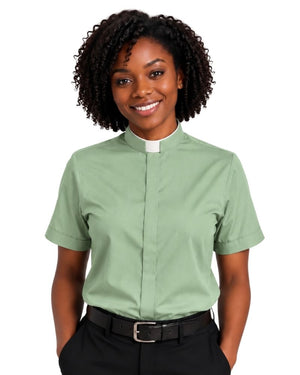 A smiling woman with curly black hair wearing a sage green short-sleeve clerical shirt with a white collar, paired with black trousers and a black belt, against a plain white background.