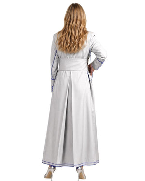 A woman viewed from the back wearing a long white or light gray cassock with blue piping trim along the sleeves, waist, and hem, standing against a plain white studio background.