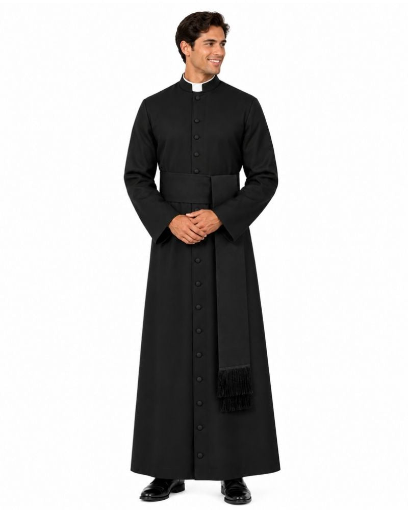A man wearing a long black clerical cassock with a high collar and a white clerical collar, standing against a plain white background.