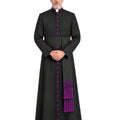 A man wearing a Black Clergy Cassock With Roman Purple Trim e with purple trim, purple buttons, and a matching purple pectoral cross, standing against a plain white background.
