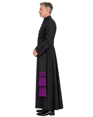 A man in profile wearing a Black Clergy Cassock With Roman Purple Trim with bright purple decorative trim, buttons, and tassels, standing against a plain white background.