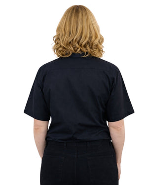 A woman viewed from the back wearing a short-sleeve black button-up  clerical shirt and black pants against a plain white background.