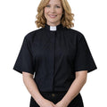 A woman with blonde hair wearing a black short-sleeve clerical shirt with a white collar, standing against a plain white background.
