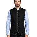 A man modeling a black waistcoat vest with white piping and white buttons, worn over a light blue dress shirt and black trousers, against a white background.