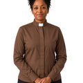 A woman wearing a brown long-sleeve clerical shirt with a white collar, paired with dark pants, standing against a plain white background.