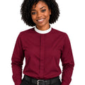 A smiling woman with curly black hair wearing a long-sleeve burgundy clerical shirt with a contrasting white crew neck collar, paired with black trousers and a black belt, against a plain white background.