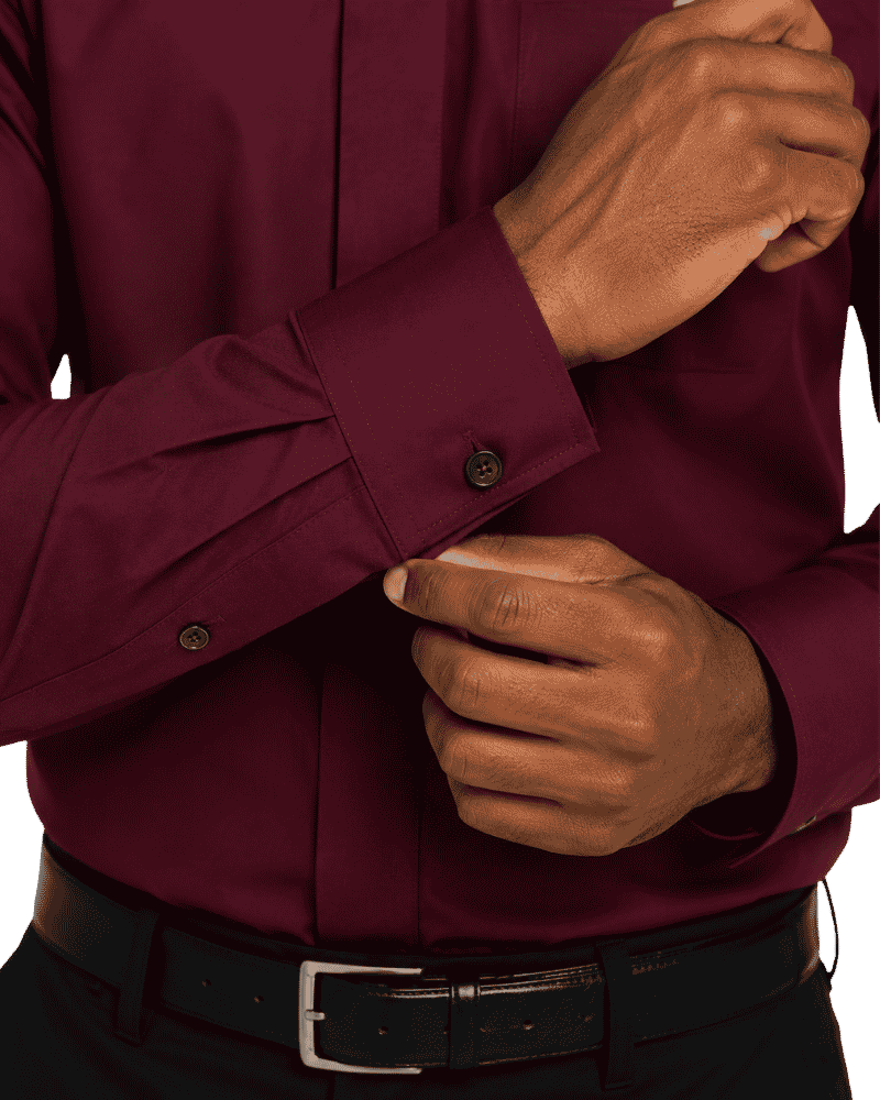 A close-up of a man's hands adjusting the cuff of a long-sleeve burgundy clerical shirt, paired with a black leather belt.