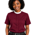 A smiling woman with curly black hair wearing a short-sleeve burgundy clerical shirt with a white collar, paired with black trousers and a black belt, against a plain white background.