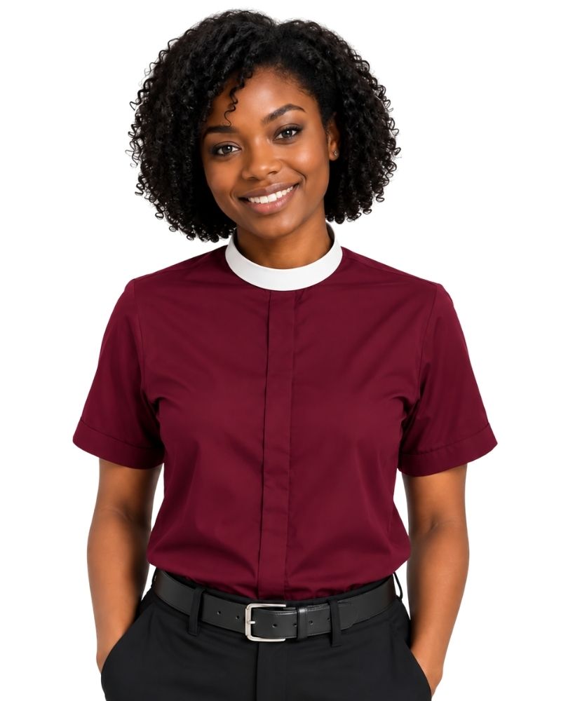 A smiling woman with curly black hair wearing a short-sleeve burgundy clerical shirt with a white collar, paired with black trousers and a black belt, against a plain white background.