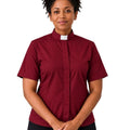 A smiling woman with curly black hair wearing a short-sleeve burgundy clerical shirt with a white collar, paired with black trousers and a black belt, against a plain white background.