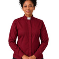 A smiling woman with curly black hair wearing a long-sleeve burgundy clerical shirt with a white collar, paired with black trousers and a black belt, against a plain white background.