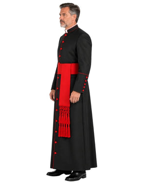 A man modeling a Classic Black Clergy Cassock with Red Trim with a wide red sash and red decorative buttons, shown in profile against a plain white background.