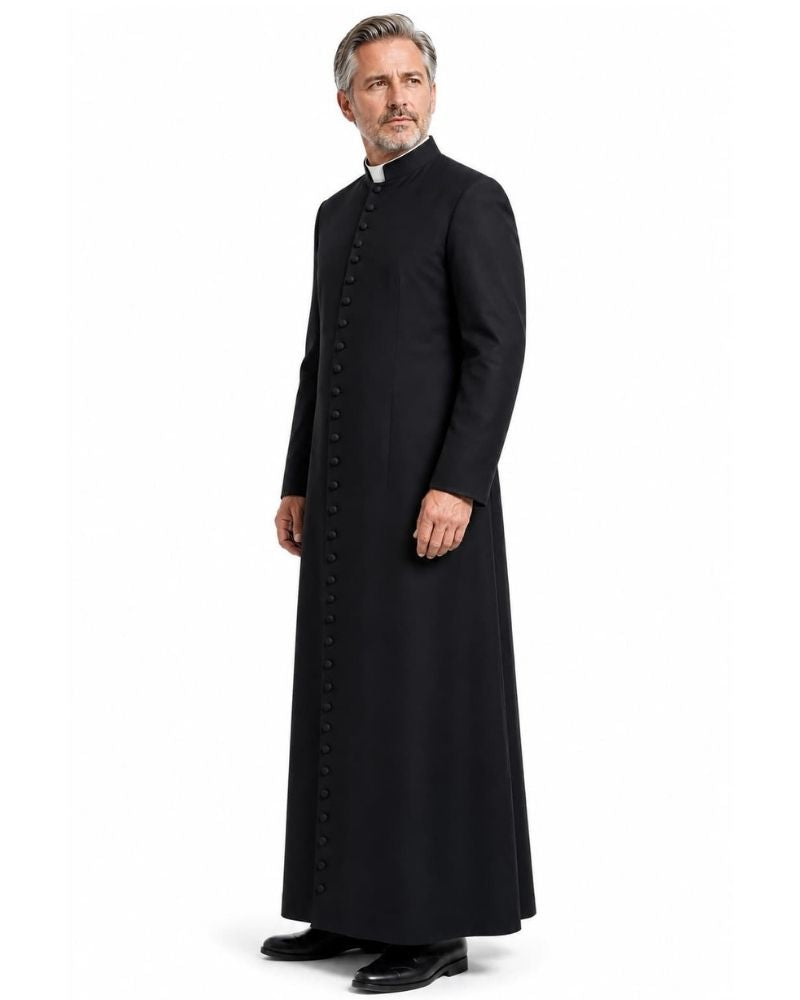 A man modeling a long black clerical Cassock with a high collar and front buttons, paired with a white collar and black dress shoes, against a plain white background.