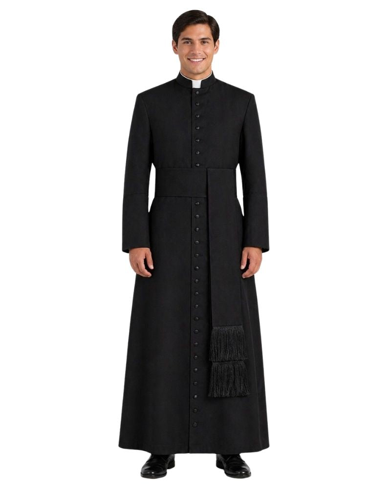 A man wearing a long black clerical cassock with a high collar, front button closure, waist tie, and a matching black scapular with fringe, photographed against a plain white background.