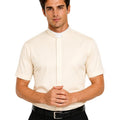 A man wearing a cream-colored short-sleeve clerical shirt with a white collar tab, paired with black trousers and a black belt, against a plain white background.