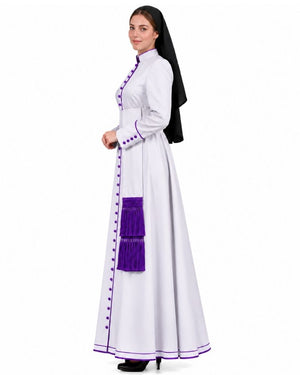 A woman modeling a white nun's habit dress with purple trim, buttons, and a purple sash, paired with a black veil, against a plain white background.