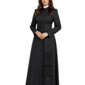 A woman modeling a long black cassock or robe with a high collar, front button placket, wide waistband, and a large fringe detail on the skirt, paired with black pointed heels against a plain white background.