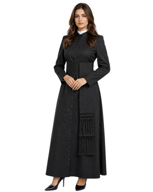 A woman modeling a long black cassock or robe with a high collar, front button placket, wide waistband, and a large fringe detail on the skirt, paired with black pointed heels against a plain white background.