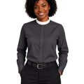 A smiling woman with curly black hair wearing a dark charcoal gray long-sleeve clerical shirt with a contrasting white crew neck collar, paired with black trousers and a black belt, against a plain white background.