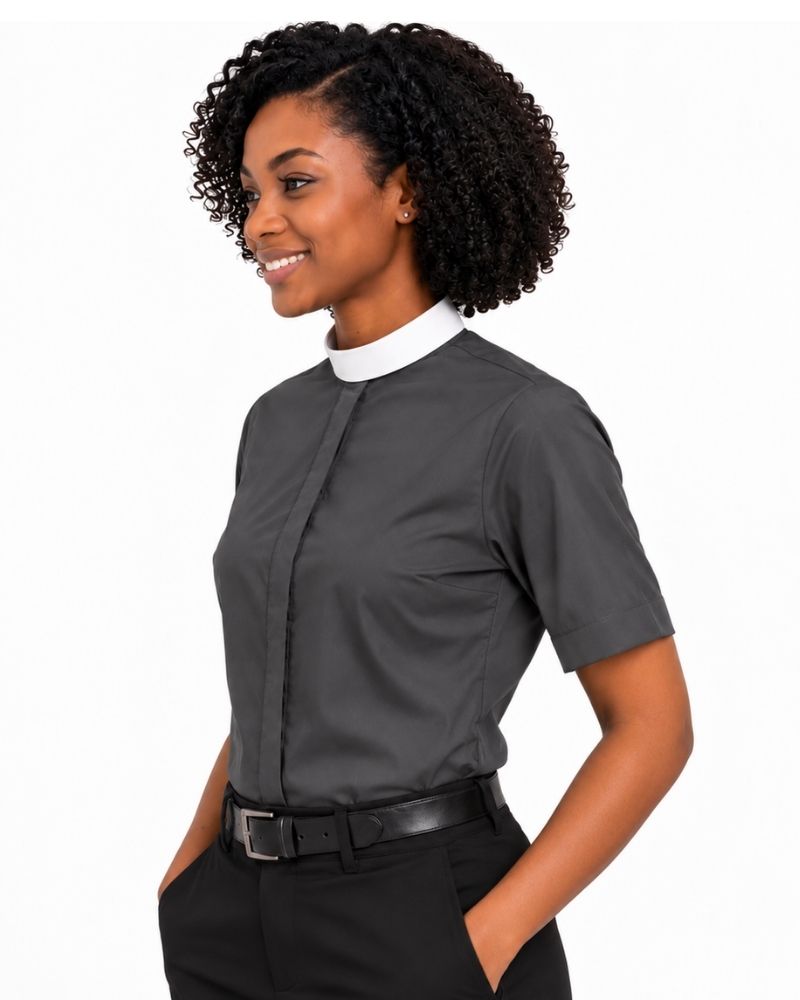 A woman with curly black hair wearing a dark gray short-sleeve clerical shirt with a white mock neck collar, paired with black trousers and a black belt, shown against a plain white background.
