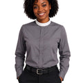 A smiling woman with curly black hair wearing a gray long-sleeve clerical shirt with a white mock neck collar, paired with black pants and a black belt, against a plain white background.