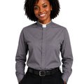 A smiling woman with curly black hair wearing a long-sleeve gray clerical shirt with a white collar, paired with black trousers and a black belt, against a plain white background.