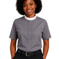 A smiling woman with curly black hair wearing a gray short-sleeve clerical shirt with a white mock neck collar, paired with black trousers and a black belt, against a plain white background.