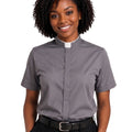A smiling woman with curly black hair wearing a short-sleeve gray clerical shirt with a white collar, paired with black trousers and a black belt, against a plain white background.