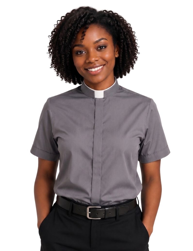 A smiling woman with curly black hair wearing a short-sleeve gray clerical shirt with a white collar, paired with black trousers and a black belt, against a plain white background.