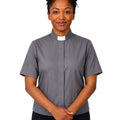 A woman wearing a short-sleeved gray clerical shirt with a white collar, standing against a plain white background.
