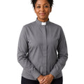 A woman wearing a gray long-sleeve clerical shirt with a white collar tab, paired with black trousers, standing against a plain white background.