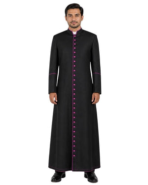 A man modeling a long black traditional Cassock with a high collar, front button closure, and magenta trim details, set against a plain white background.