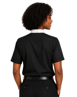 A woman with curly black hair is shown in profile wearing a black short-sleeve  clerical shirt with a contrasting white high collar, paired with black trousers and a black belt, against a plain white background.