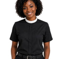 A smiling woman with curly black hair wearing a black short-sleeve clerical shirt with a white collar, paired with black trousers and a black belt, against a plain white background.