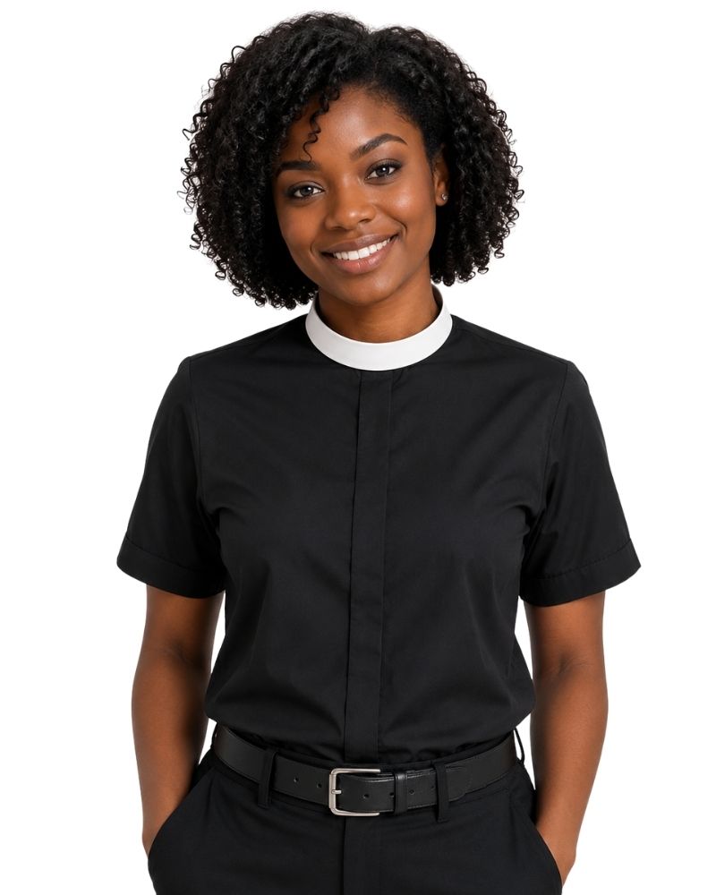 A smiling woman with curly black hair wearing a black short-sleeve clerical shirt with a white collar, paired with black trousers and a black belt, against a plain white background.