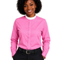 A smiling woman with curly black hair wearing a bright pink long-sleeve clerical shirt, black trousers, and a black belt, photographed against a plain white background.