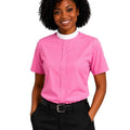 A smiling woman with curly black hair wearing a bright pink short-sleeve uniform shirt with a white mock neck collar, paired with black trousers and a black belt, against a plain white background.