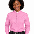 A smiling woman with curly black hair wearing a pink long-sleeve clerical shirt with a white collar, paired with black trousers and a black belt, against a plain white background.