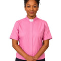 A woman wearing a short-sleeve pink clerical shirt with a white collar, paired with black trousers, standing against a plain white background.
