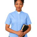 A woman with short curly black hair wearing a light blue short-sleeve clerical shirt with a white collar, holding a black book, set against a plain white background.